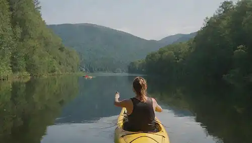 3 kayaks, una persona rematando uno con un perro de pie en la espalda, otro kayak a distancia con la onda de la persona, y tercer kayak con persona dando pulgares hacia arriba, cielo soleado, lago tranquilo, exuberante vegetación, ambiente tranquilo, vibraciones de verano