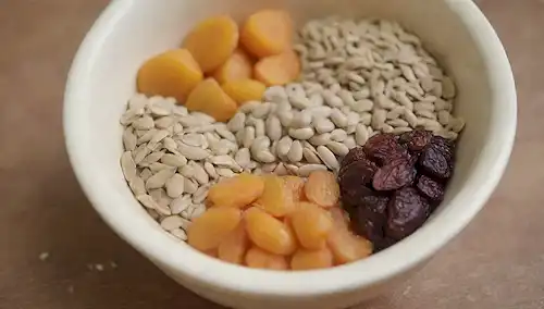 Un primer plano de un tazón lleno de frutas y semillas secas surtidas, como pasas, albaricoques, semillas de calabaza y semillas de girasol, con un poco de heno fresco o hierba cerca
